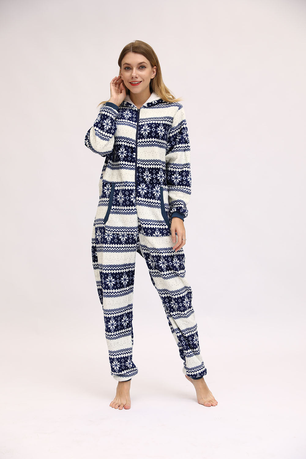 Full-length angled view of a woman wearing the Noelle navy blue and white Christmas pajama onesie, featuring horizontal rows of navy reindeer, snowflake designs, geometric winter patterns, a zippered front, fitted-looking hood, long sleeves with elasticized cuffs, and matching full-length pajama legs gathered at the ankles.