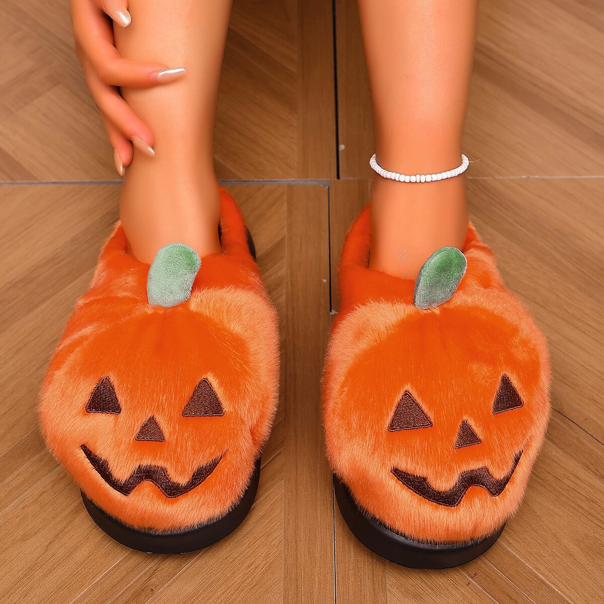 Orange women’s jack-o’-lantern plush slippers shown worn from the front, featuring rounded fuzzy pumpkin uppers, embroidered black smiling Halloween faces, green stem accents, open-back styling, and thick black soles for a cozy spooky-season look.