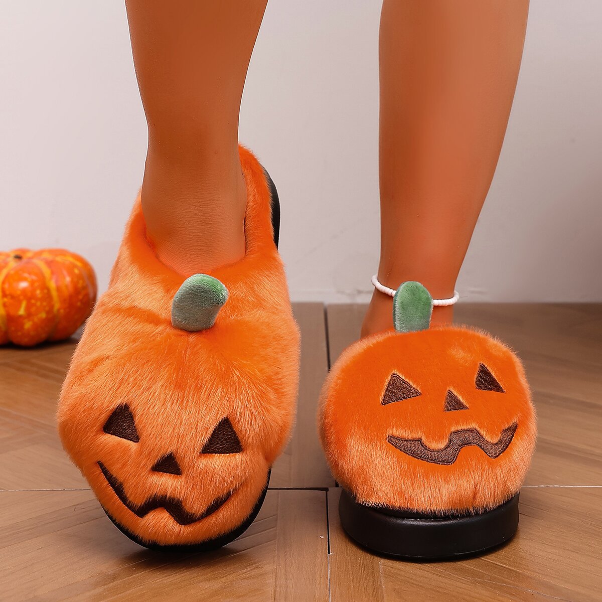 Orange pumpkin Halloween house slippers for women shown from a slightly elevated front view, featuring soft plush pumpkin-shaped uppers, black embroidered jack-o’-lantern faces, small green stems, open-back design, and chunky black soles clearly visible beneath both slippers.