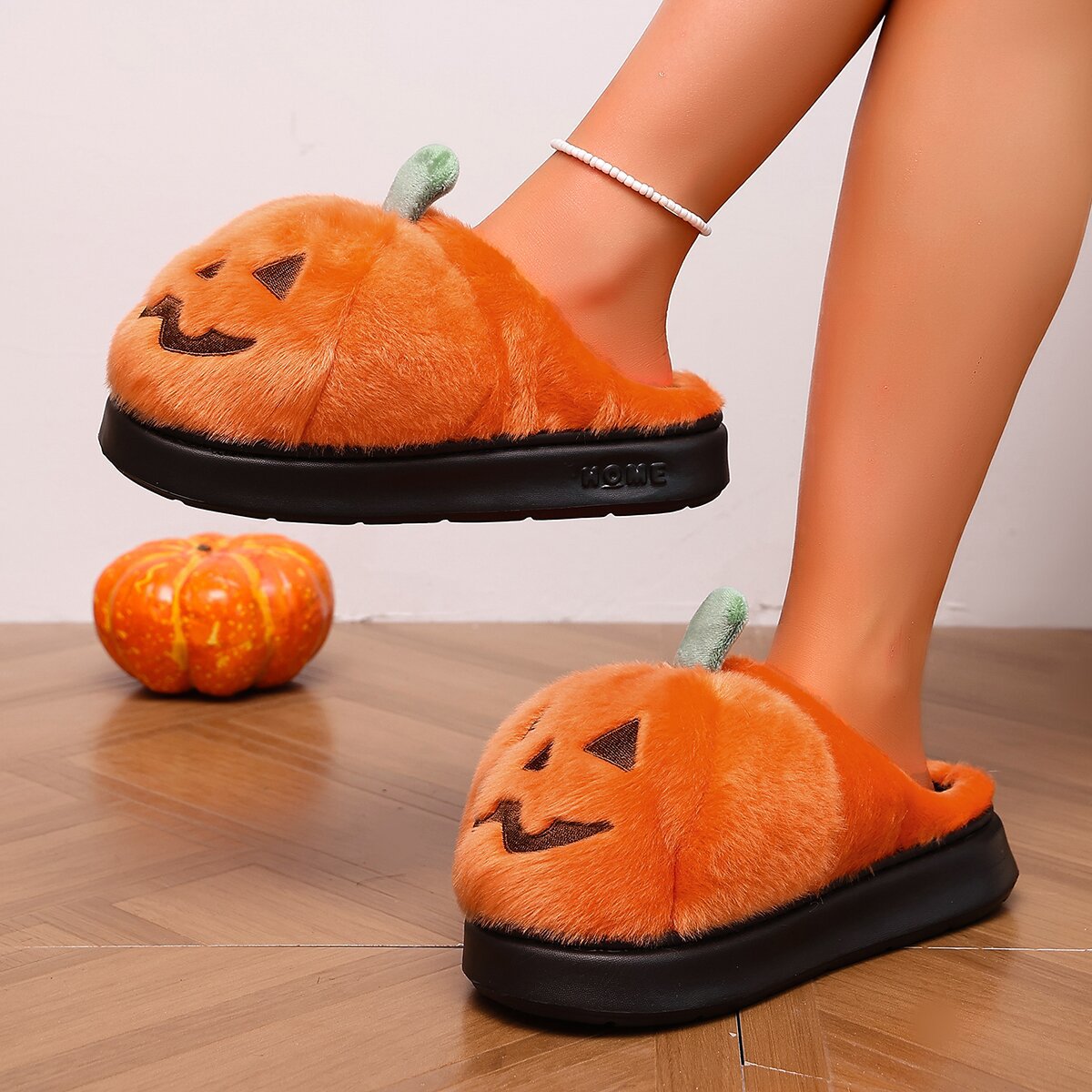 Orange women’s plush Halloween slippers designed like fuzzy pumpkins with embroidered black jack-o’-lantern faces, small green stems, open-back construction, and thick black soles, shown worn from a side angle with one foot lifted beside decorative pumpkins for a festive fall lifestyle view.