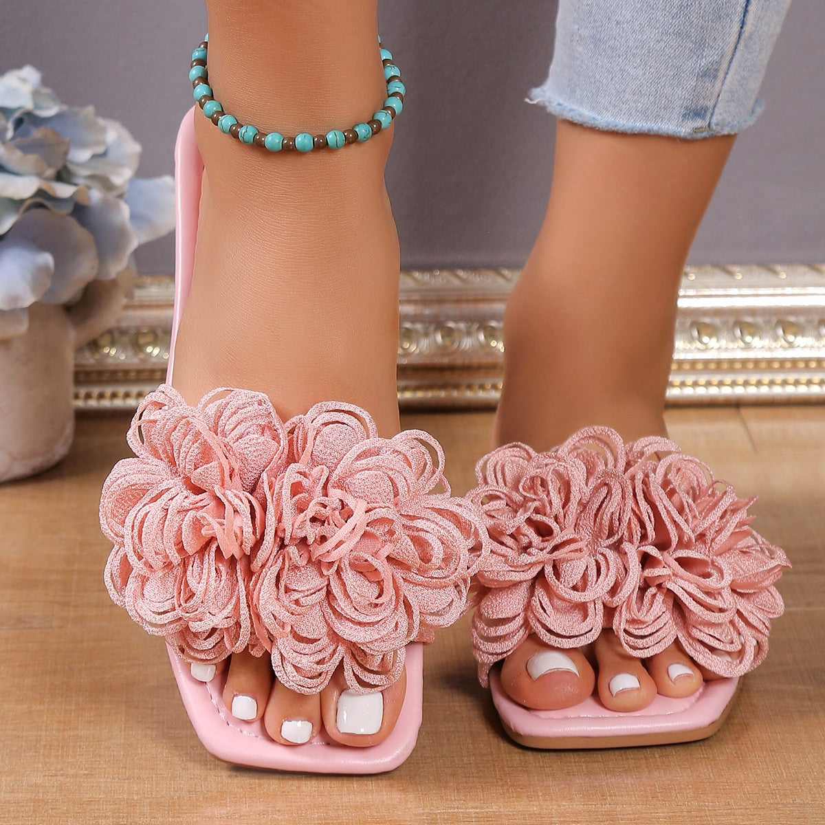 Pink women’s flat slide sandals featuring oversized layered 3D floral embellishments across the wide upper strap, an open-toe design, and flat slip-on silhouette, shown worn from the front with both feet visible for a clear view of the dimensional flower detailing.