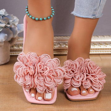 Pink women’s flat slide sandals featuring oversized layered 3D floral embellishments across the wide upper strap, an open-toe design, and flat slip-on silhouette, shown worn from the front with both feet visible for a clear view of the dimensional flower detailing.