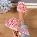 Pink women’s floral flat slide sandals featuring layered 3D flower appliqués across the upper strap, an open-toe design, and slim flat sole, shown from a side angle to highlight the raised floral embellishments and feminine slip-on silhouette.