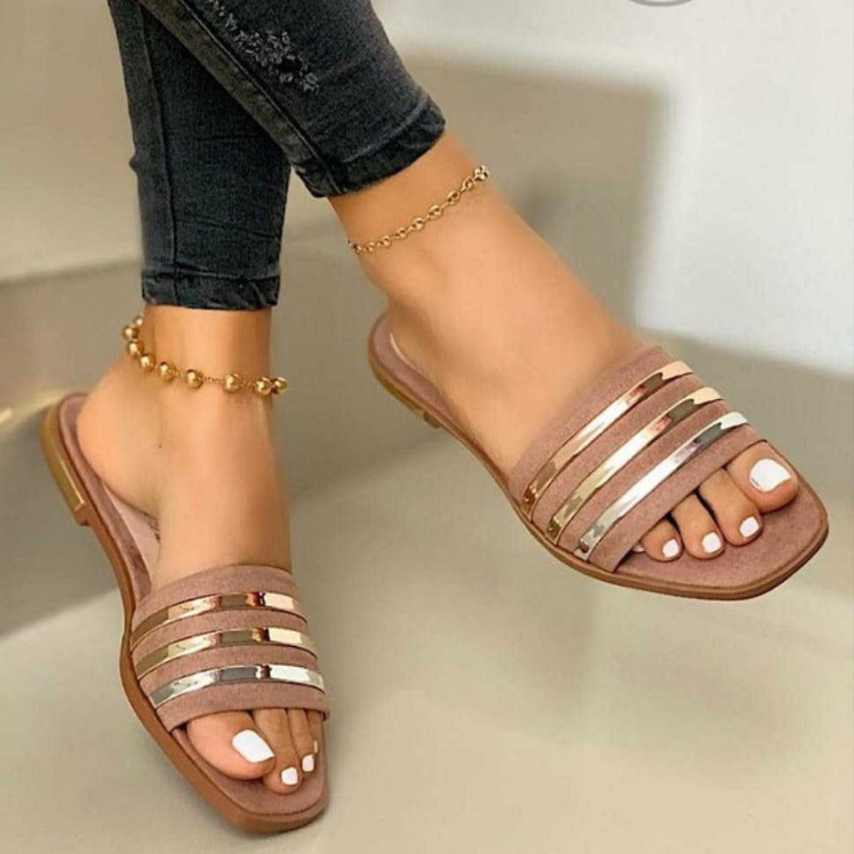 Pink women’s flat slide sandals featuring layered horizontal pink straps with shiny metallic accents, an open-toe design, slim low-profile flat sole, and slip-on construction, shown worn on both feet from an angled front view for a clear look at the soft pink color and reflective stripe detailing.