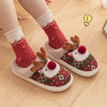 Red and green women’s reindeer Christmas plush slippers shown worn from the front, featuring raised 3D reindeer decorations with brown antlers and red pom-pom noses, festive patterned uppers, beige plush trim, and cozy open-back styling paired with red socks.