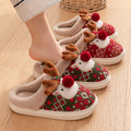 Women’s red and green reindeer holiday plush slippers shown worn from an angled side view, highlighting the 3D reindeer faces, brown antlers, red pom-pom noses, festive patterned fabric, beige fuzzy lining, and cozy slip-on construction for Christmas lounging.