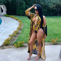 Safiya women's black-and-gold printed resort swimwear set shown on a model wearing a plunging black swimsuit decorated with elaborate gold baroque-style floral and scroll patterns, paired with a long matching open-front cover-up featuring coordinated black-and-gold ornamental designs.