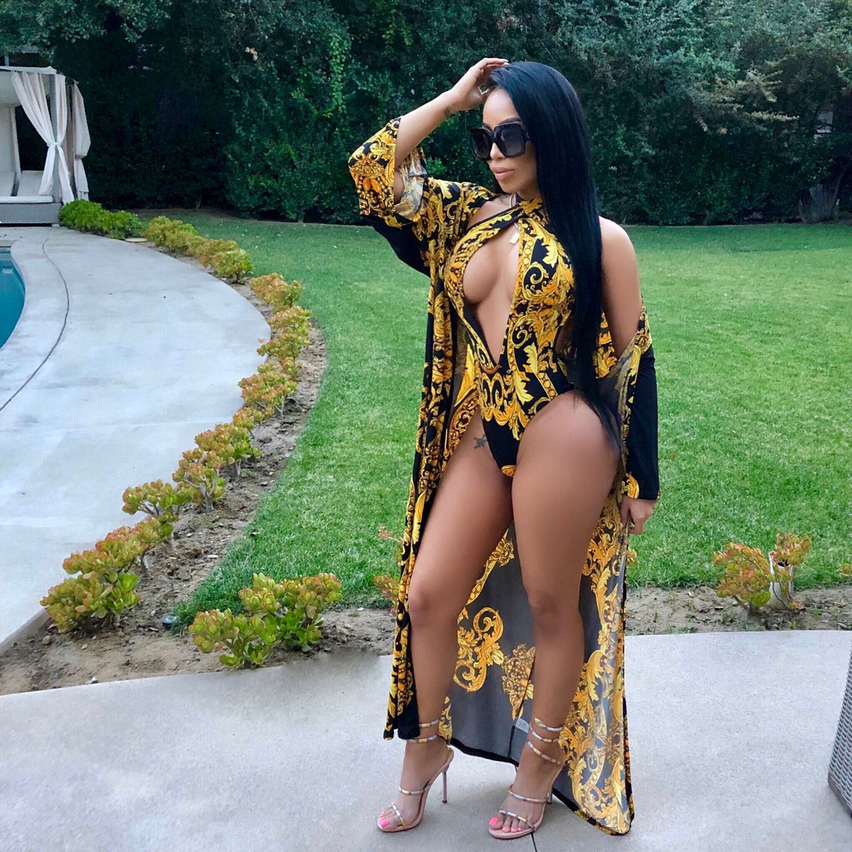 Safiya women's black-and-gold printed resort swimwear set shown on a model wearing a plunging black swimsuit decorated with elaborate gold baroque-style floral and scroll patterns, paired with a long matching open-front cover-up featuring coordinated black-and-gold ornamental designs.