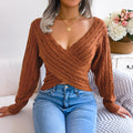 Woman seated on a bed wearing a cinnamon brown V-neck twist-front cropped knit sweater with blue jeans.