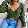 Woman seated indoors wearing an olive green twist-front V-neck cropped knit sweater with high-waisted blue jeans.