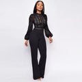 Seraphina women's elegant black evening jumpsuit shown from the front, featuring an intricate semi-sheer lace bodice with floral and geometric patterns, high scalloped neckline, long sheer black sleeves with gathered cuffs, fitted waist, and flowing black trousers.