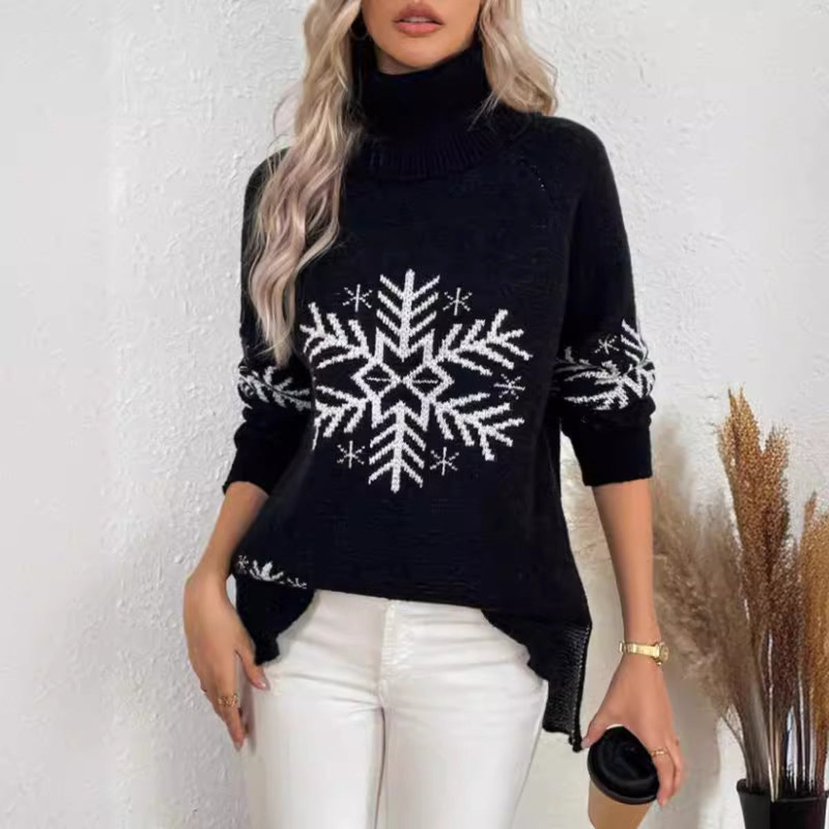 Woman wearing a Black Snowfall Luxe Turtleneck Holiday Sweater with a large white snowflake design, white pants, and a small black handbag.
