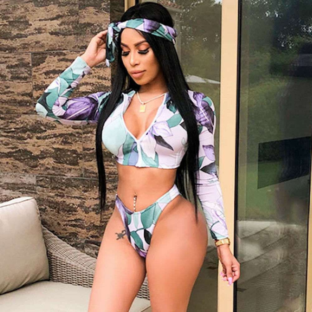 Front view of a model wearing a long-sleeve cropped zip-front bikini top and coordinating bottoms with lavender flowers, teal-green leaves, and a white tropical background.