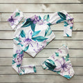 Close-up front view of the Soleil women's cropped long-sleeve swim top showing a central zipper, plunging V-shaped opening, lavender floral motifs, teal-green tropical leaves, and a fitted cropped hem ending above the waist.