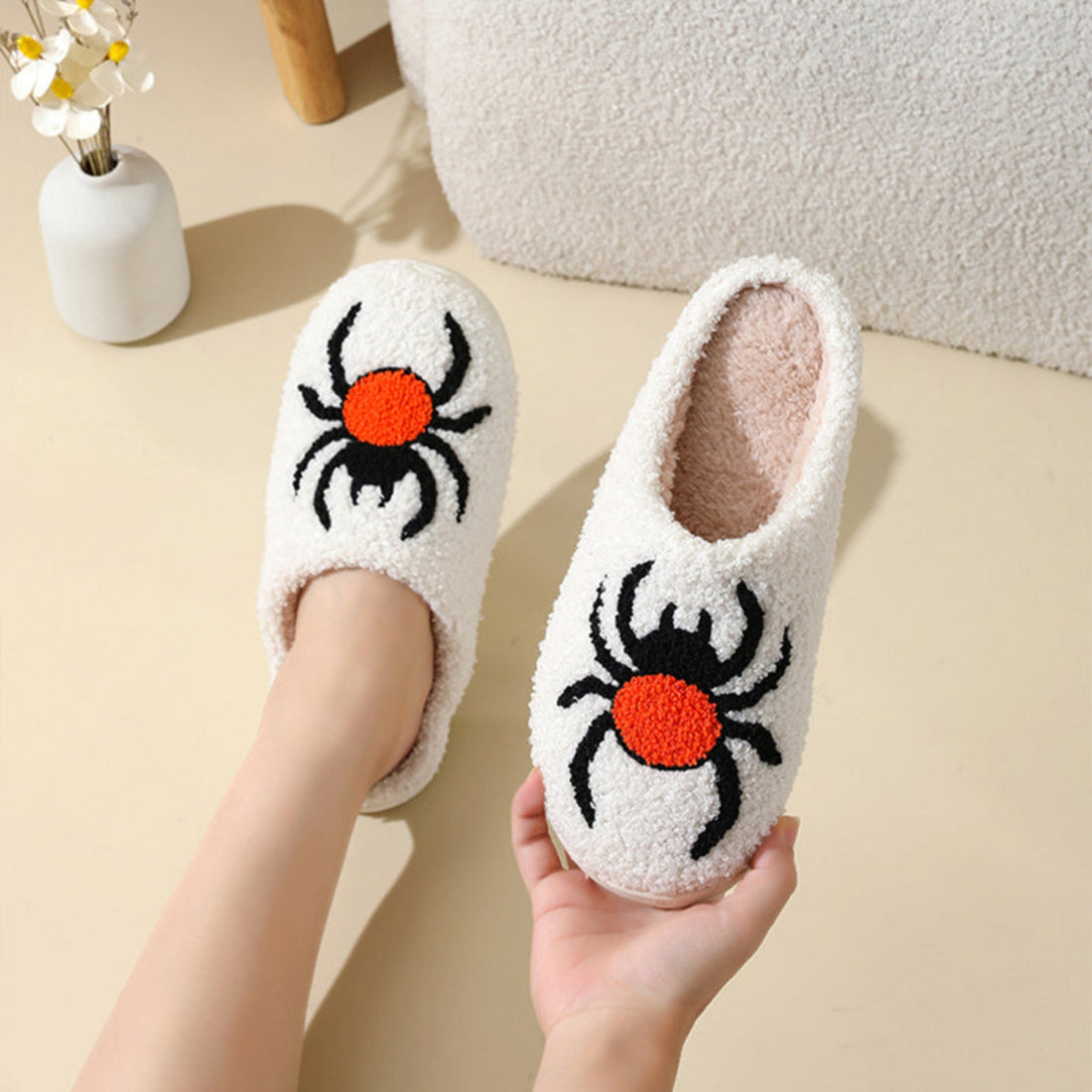 Cream women’s plush Halloween slippers featuring large embroidered black spiders with bright orange-red bodies across the front, a soft fuzzy closed-toe construction, cozy cream lining, and rounded slip-on silhouette, displayed together from the front for a clear view of the bold spider artwork and plush texture.