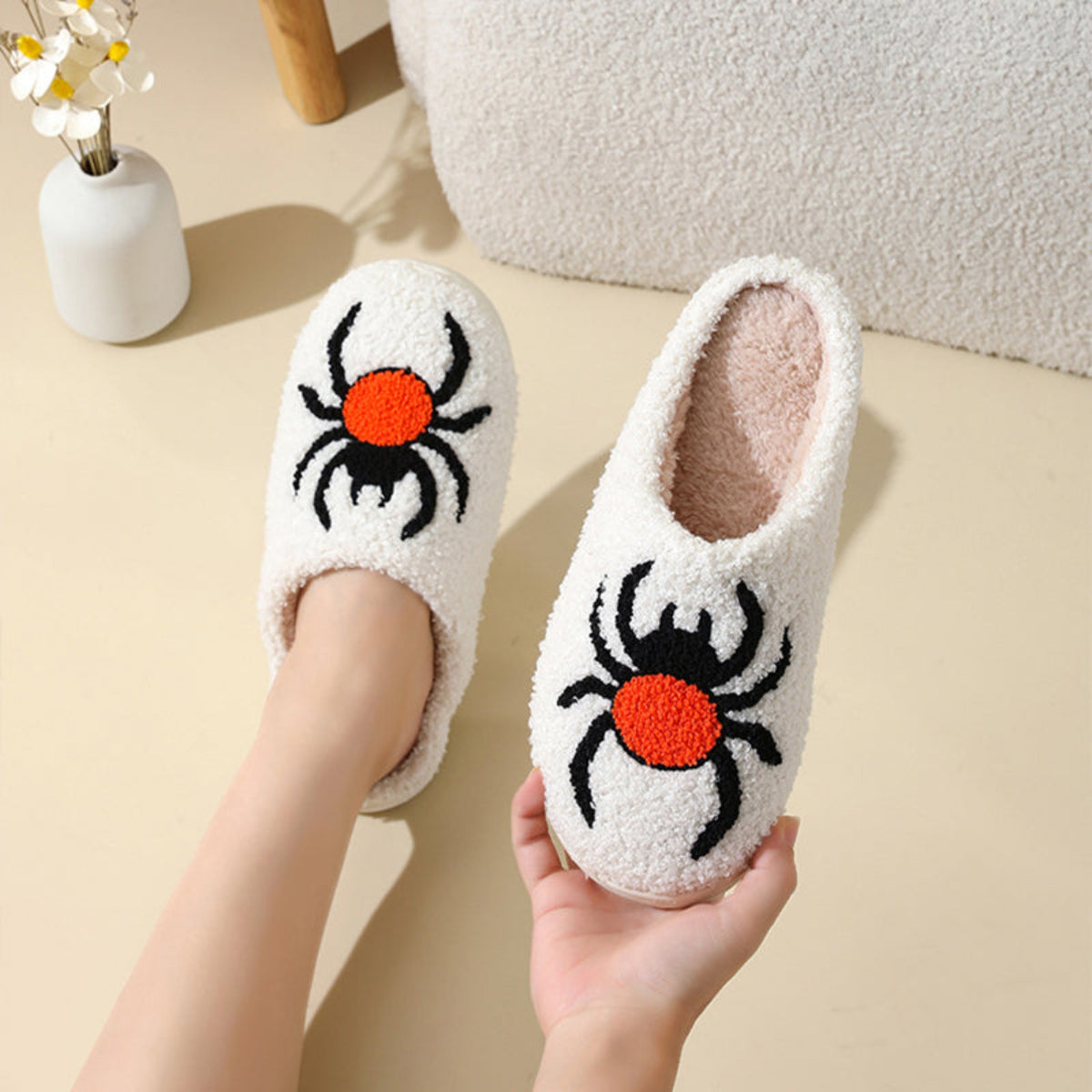 Cream women’s Halloween plush slippers featuring oversized black spider designs with bright orange-red centers, soft fuzzy closed-toe uppers, and cozy slip-on construction, shown worn and held from above to highlight the spider embroidery, rounded shape, and seasonal Halloween design.