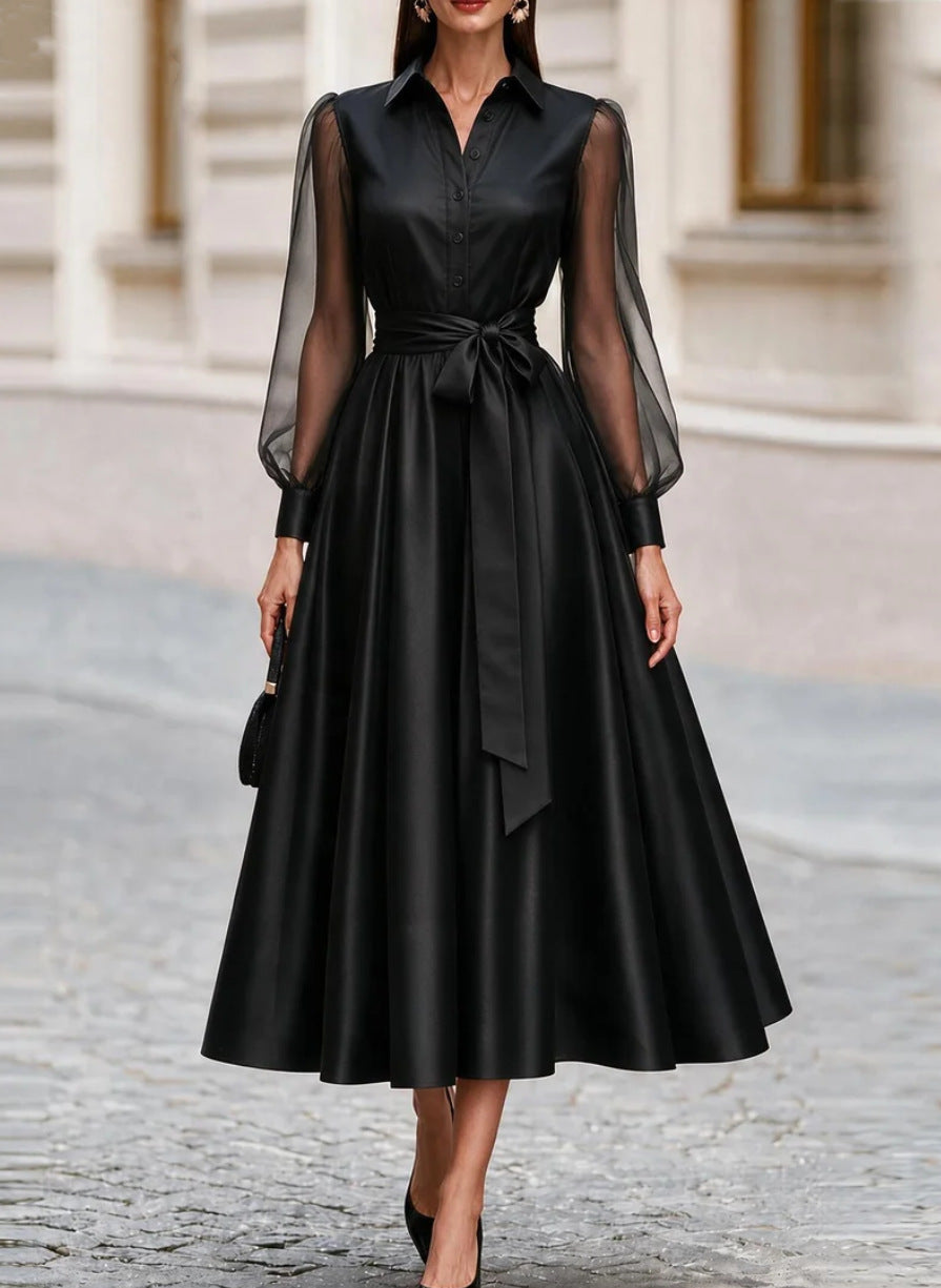 A woman wearing a long-sleeve satin midi dress in deep burgundy with sheer mesh sleeves and a matching waist tie, standing on a cobblestone street.