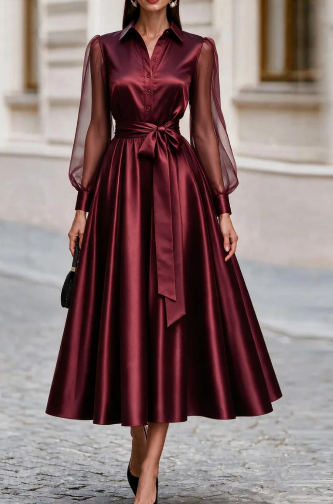 A woman wearing a long-sleeve satin midi dress in deep burgundy with sheer mesh sleeves and a matching waist tie, standing on a cobblestone street.
