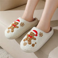 White women’s gingerbread holiday slippers shown worn from an angled front view, featuring plush fuzzy uppers, embroidered gingerbread figures with red Christmas details, brown snowflakes, rounded closed toes, and cozy slip-on styling for seasonal indoor wear.