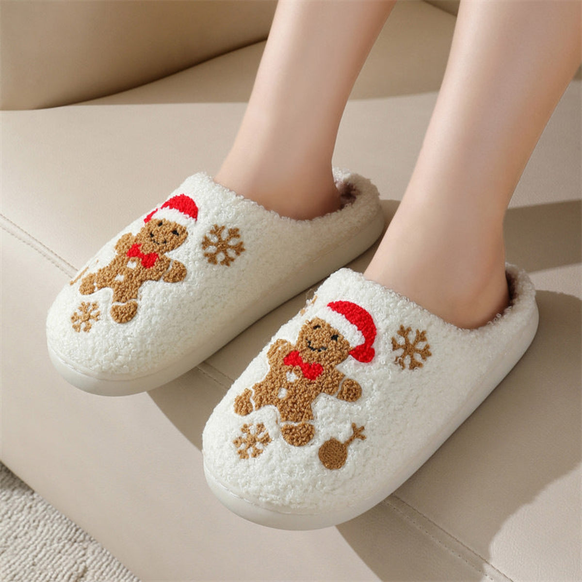 White women’s gingerbread holiday slippers shown worn from an angled front view, featuring plush fuzzy uppers, embroidered gingerbread figures with red Christmas details, brown snowflakes, rounded closed toes, and cozy slip-on styling for seasonal indoor wear.
