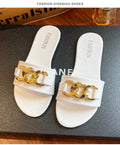 White women's flat slide sandals featuring a wide white strap, oversized shiny gold-tone chain buckle detail, open-toe silhouette, and flat white footbed, displayed as a matching pair on a dark textured surface.