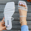 White women’s minimalist flat sandals featuring multiple slim rounded straps across the toes and midfoot, an open square-toe silhouette, low flat sole, and clean modern design, shown with one sandal worn on the foot and the matching sandal held beside it for a detailed top view.