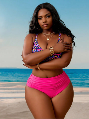 Zuri women's two-piece swimsuit modeled on a sandy beach with the ocean in the background, featuring a vibrant multicolor leopard-print bikini top with pink, purple, blue, and yellow accents, narrow shoulder straps, and bright hot-pink high-waisted bikini bottoms with gathered ruching across the waist. The model has long black wavy hair and poses with her arms crossed over her chest.