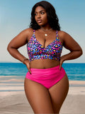 Full front view of the Zuri women's high-waisted two-piece swimsuit worn by a curvy model standing on a tropical beach, featuring a colorful leopard-print V-neck bikini top with wide shoulder straps and a gathered center-front detail, paired with solid hot-pink high-waisted bottoms featuring flattering ruching across the midsection and full front coverage.